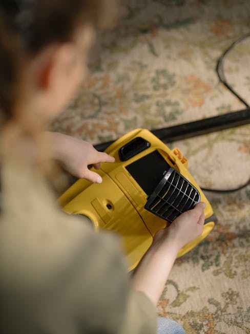 A person kneeling on a patterned area rug inside a room is cleaning a yellow and black vacuum cleaner, inserting a cloth into the dirt chamber for surface cleaning. The room features wooden flooring,
